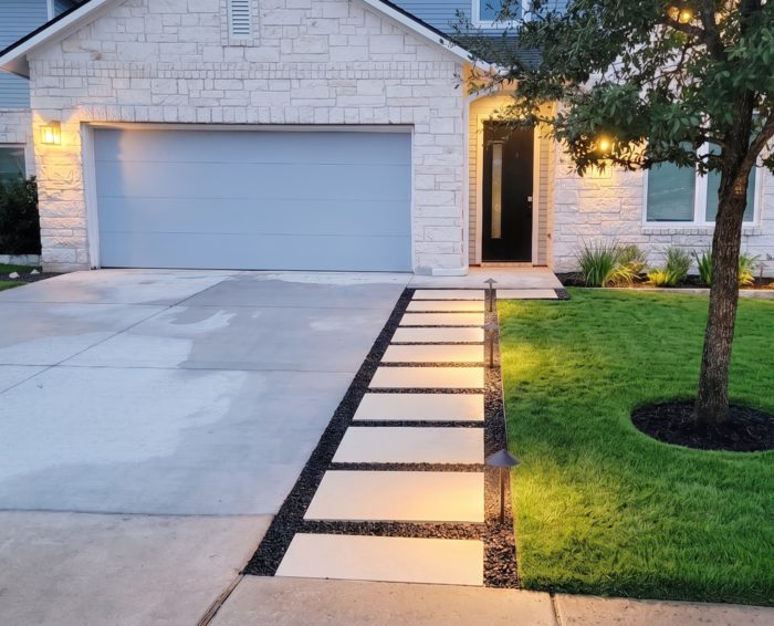 Front yard landscaping with a lit paver walkway, fresh sod and a mulched tree ring at dusk